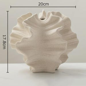 Op de afbeelding: Een cr&egrave;mekleurige, getextureerde keramische vaas met een uniek, gerimpeld ontwerp. De vaas is ongeveer 17,8 cm hoog en 20 cm breed. De opening is klein en de algehele vorm is organisch en sculpturaal. De vaas is een decoratief woonaccessoire.