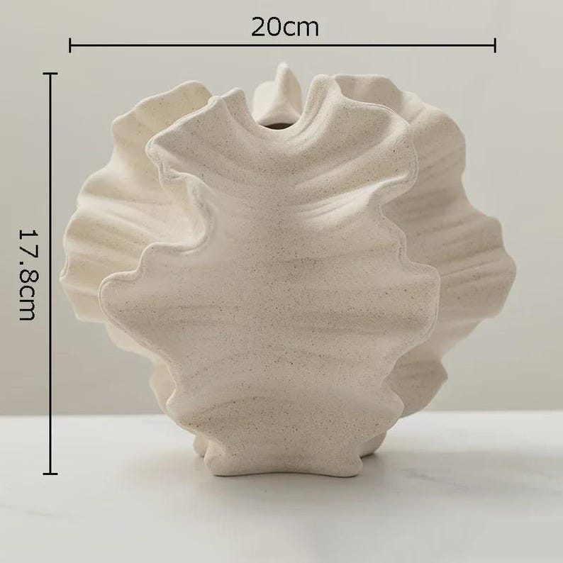 Op de afbeelding: Een cr&egrave;mekleurige, getextureerde keramische vaas met een uniek, gerimpeld ontwerp. De vaas is ongeveer 17,8 cm hoog en 20 cm breed. De opening is klein en de algehele vorm is organisch en sculpturaal. De vaas is een decoratief woonaccessoire.
