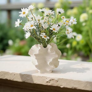 Op de afbeelding: Een cr&egrave;mekleurige, gerimpelde vaas bevat een boeket witte madeliefjes met gele centra. De vaas heeft een uniek, gestructureerd ontwerp. De bloemen zijn gerangschikt in een natuurlijke, organische stijl, wat een frisse en uitnodigende presentatie cre&euml;ert.