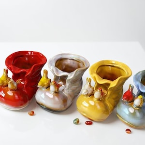 May include: A collection of four ceramic boot-shaped planters in red, gray, yellow, and blue. Each planter features a decorative cluster of small bird figurines. The planters are glossy and have a smooth finish.
