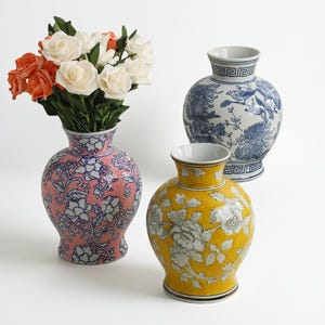 May include: Three decorative ceramic vases with floral designs. One pink vase holds orange and white flowers. A blue and white vase features a bird and floral pattern. A yellow vase displays white floral motifs.