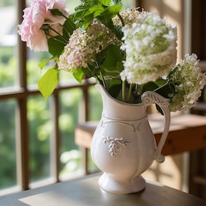 May include: A white ceramic pitcher with a decorative handle and embossed floral design, filled with pink and white flowers and green foliage. The pitcher is sitting on a wooden surface near a window.