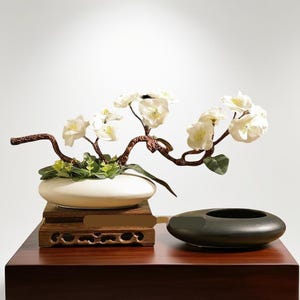 Könnte beinhalten: Dekorative Blumengesteck mit weißen Blüten auf einem braunen Zweig, in einem ovalen weißen Keramiktopf. Der Topf steht auf einem geschnitzten Holzsockel. Eine dunkelgraue ovale Keramikschale daneben.