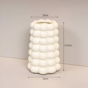 May include: A white ceramic vase with a unique bubble design. The vase is 6.9 inches tall, 4 inches wide at the top, and 4.7 inches wide at the base. The vase has a wavy top edge and a smooth, glossy finish. Home decor.