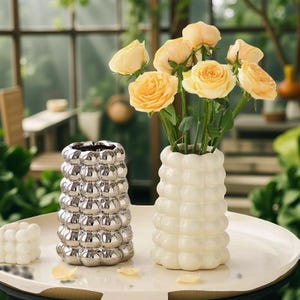 May include: Two decorative vases, one silver and one white, sit on a white table. The silver vase has a bubble design, while the white vase holds yellow roses. The vases are in front of a window.