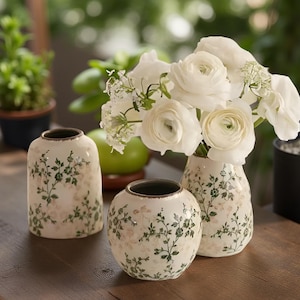 Puede incluir: Tres jarrones de cerámica con motivos florales. Los jarrones son blancos con diseños florales verdes y rosas. Uno es cilíndrico, otro redondo y el tercero es más pequeño. Los jarrones están llenos de flores blancas.