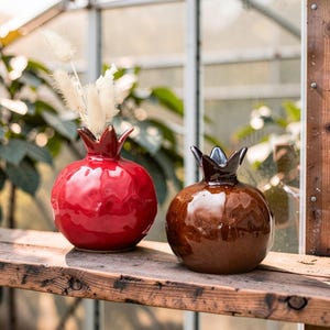 May include: Two ceramic pomegranate-shaped vases, one red and one brown, sit on a wooden shelf. The red vase holds dried white flowers. Both vases have a glossy finish and crown-like tops, adding a decorative touch to any space.