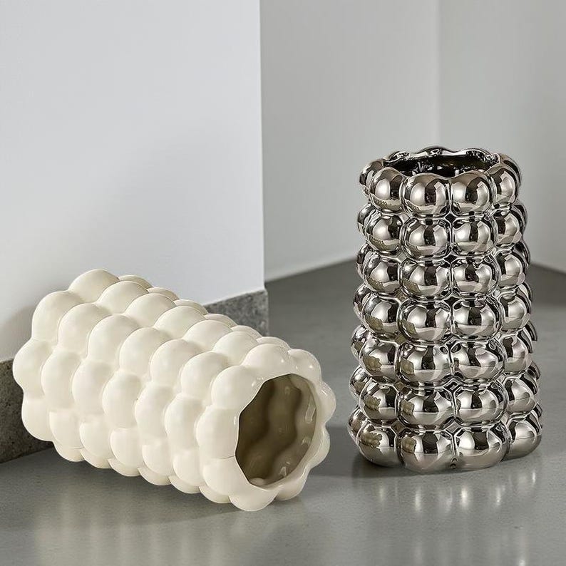 May include: Two decorative vases. One is a cylindrical, off-white vase with a bubble-like texture. The other is a silver-colored, cylindrical vase with a similar bubble design. Both vases are on a gray surface.
