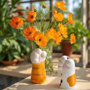 May include: Two cat paw-shaped vases, one orange and white, the other white and orange, hold vibrant orange flowers. The vases are on a wooden surface, with a blurred green background. The flowers are in full bloom.