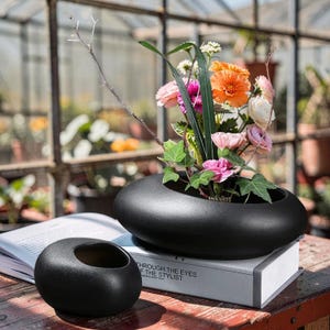 Puede incluir: Un jarrón de cerámica negro, de forma ovalada, contiene un vibrante arreglo floral con flores naranjas, rosas y blancas, acentuado por follaje verde. Un jarrón negro más pequeño y redondo está cerca. El arreglo está sobre un libro.