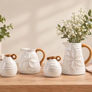 Può includere: Un set di quattro vasi in ceramica con smalto bianco maculato e dettagli marroni. Due piccoli vasi rotondi hanno manici laterali, mentre due brocche presentano motivi floreali in rilievo e manici marroni. Una brocca contiene fiori bianchi.