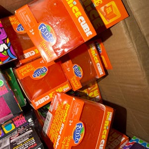 May include: A box filled with orange Nee Doh Nice Cube toys. Each package features the Fanta logo and the words "Stupid Squish!" and "Nee Doh Nice Cube!" The toys are recommended for ages 3+.