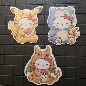 Pode incluir: Tr&ecirc;s autocolantes hologr&aacute;ficos com Hello Kitty vestida como personagens Pok&eacute;mon. Um &eacute; Pikachu, outro &eacute; Snorlax com um Pikachu a dormir, e o terceiro &eacute; Charizard. Cada autocolante segura uma Pok&eacute; Bola e tem um la&ccedil;o vermelho.
