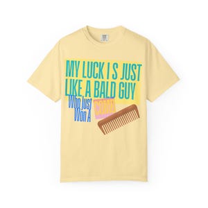 May include: A light yellow t-shirt with the text "MY LUCK IS JUST LIKE A BALD GUY Who Just Won A COMB" in blue, teal, and pink. A brown comb graphic is also featured on the shirt.