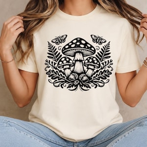 Könnte beinhalten: Ein cremefarbenes Kurzarm-T-Shirt mit einem auffälligen schwarzen Grafikdruck auf der Vorderseite. Das Design zeigt drei Pilze, umgeben von Farnblättern, kleinen Eicheln und zwei Motten, die sich in den oberen Ecken der Illustration befinden.