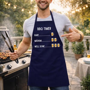 May include: A navy blue apron with the text "BBQ TIMER" and beer mug illustrations indicating levels of doneness: rare, medium, and well done. A person points at the apron near a barbecue.