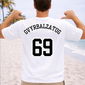 Può includere: T-shirt bianca con la scritta "GVYRBALZATUG" e il numero "69" in grassetto nero. La maglietta è semplice sul davanti. L'immagine mostra il retro della maglietta, indossata su una spiaggia.