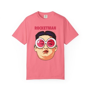May include: Pink t-shirt with the word "ROCKETMAN" printed across the top. The shirt features a graphic of a person wearing large, pink-tinted sunglasses. The person has dark hair and a neutral expression.