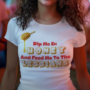 May include: White t-shirt featuring the text "Dip Me In Honey And Feed Me To The Lesbians" in red and yellow. A cartoon honey dipper with dripping honey is above the text.