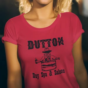 Könnte beinhalten: Ein rotes T-Shirt mit dem Wort "DUTTON" in einer kräftigen Schrift im Western-Stil. Darunter befindet sich eine Grafik einer Person in einem Fass und der Text "Day Spa & Saloon". Das Shirt hat kurze Ärmel und einen Rundhalsausschnitt.