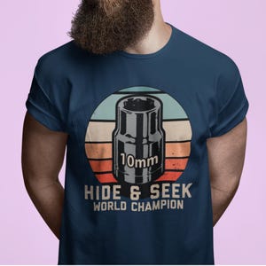 Könnte beinhalten: Marineblaues T-Shirt mit einer Grafik eines 10-mm-Steckschlüssels in einem Kreis mit Retro-Sonnenuntergangsdesign. Der Text "HIDE & SEEK WORLD CHAMPION" ist darunter gedruckt.