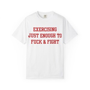 Puede incluir: Camiseta blanca con el texto rojo "EXERCISING JUST ENOUGH TO FUCK & FIGHT". La camiseta tiene cuello redondo y mangas cortas. El texto está en negrita, con fuente sans-serif.