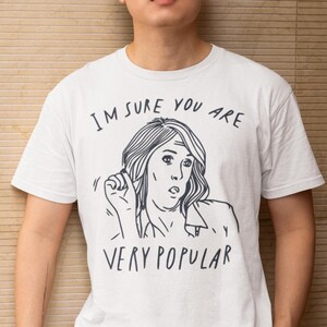 以下が含まれることがあります： 「I'M SURE YOU ARE VERY POPULAR」の文字と、驚いた表情の女性の白黒イラストがプリントされた白いTシャツ。半袖でクルーネックのデザインです。