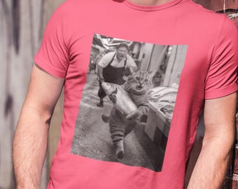 May include: A pink t-shirt featuring a black and white image of a cat running with a fish, pursued by a man. The cat is the focal point, with the man and fish market in the background.