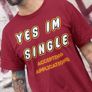 May include: A maroon t-shirt with the text "YES IM SINGLE" in white letters with a yellow outline. Below the text are the words "ACCEPTING APPLICATIONS" in orange.