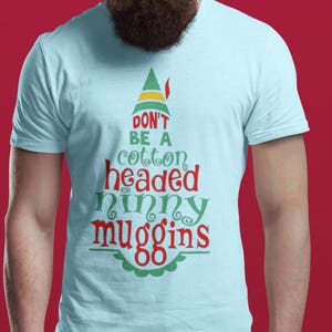 Pode incluir: Camiseta azul clara com o texto "DON'T BE A cotton headed ninny muggins" em vermelho e verde. O design inclui um gráfico de chapéu de elfo. A camisa é de manga curta.