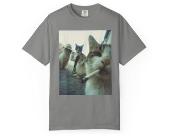 May include: A gray t-shirt featuring a humorous image of four cats. One cat is smoking a cigarette, while the others are looking at the camera. The cats are wearing a cowboy hat and looking at the camera.