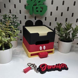 May include: A Mickey Mouse-themed desk organiser in red, black, and yellow, with a matching keychain that says "Jennifer". A green Chinese character decorates the background. The organiser has a drawer and a notepad.