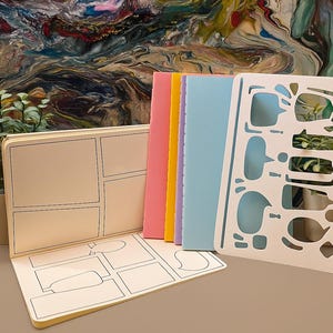 Könnte beinhalten: Ein offenes Comicbuch mit leeren Panels und einer Sprechblasen-Schablone. Daneben bunte Notizbücher in Rosa, Gelb und Blau. Auf beiden Seiten der Artikel befinden sich kleine Topfpflanzen.