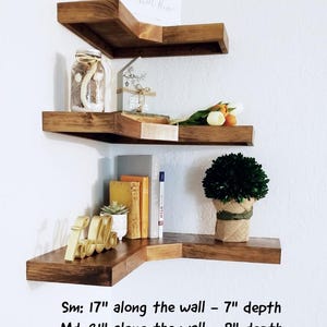 May include: Three wooden corner shelves in a warm brown finish are mounted on a white wall. The shelves are decorated with a variety of items, including books, plants, and decorative objects. Text on the image reads: Sm: 17" along the wall - 7" depth, Md: 21" along the wall - 8" depth, Lg: 25" along the wall - 9" depth.