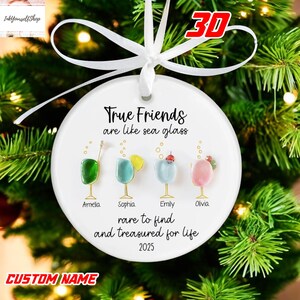A white ornament with a ribbon, featuring illustrations of four different colored drinks and the text "True Friends are like sea glass" along with the names "Armelia, Sophia, Emily, Olivia" and the year "2025".