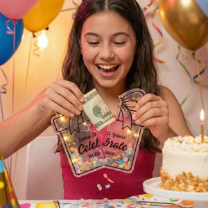 K&ouml;nnte beinhalten: Ein junger Mensch &ouml;ffnet ein Geburtstagsgeschenk, das einen 20-Dollar-Schein enth&auml;lt. Das Geschenk ist ein durchsichtiger Beh&auml;lter mit Konfetti und Lichterketten, auf dem die Worte "Today Is Yours Celebrate With Love" stehen. Ein Geburtstagskuchen und Luftballons sind im Hintergrund zu sehen.