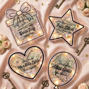 Puede incluir: Una colecci&oacute;n de adornos con forma de regalo llenos de moneda estadounidense, confeti y luces de hadas. Las formas incluyen una caja de regalo, una estrella, un coraz&oacute;n y un globo, cada uno con texto en alem&aacute;n. Llaves antiguas y rosas est&aacute;n dispersas.