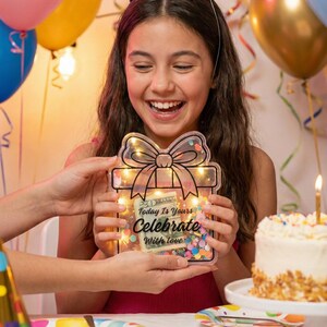 K&ouml;nnte beinhalten: Eine l&auml;chelnde Person h&auml;lt eine transparente Geschenkbox mit Schleifendesign, gef&uuml;llt mit Konfetti und Lichtern. Auf der Box steht der Text "Today Is Yours Celebrate With love". Ein Geburtstagskuchen und Luftballons sind im Hintergrund.