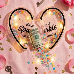K&ouml;nnte beinhalten: Auf rosa Hintergrund zwei herzf&ouml;rmige, transparente Beh&auml;lter mit dem Text "Born to Sparkle" und "Keep shining". Die Beh&auml;lter sind mit Konfetti und Lichterketten gef&uuml;llt. Eine aufgerollte Banknote liegt dazwischen, mit verstreutem Konfetti, Schl&uuml;sseln, Rosenbl&auml;ttern und goldenen Sternen.