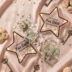 Puede incluir: Dos cajas de regalo en forma de estrella con el texto "Ein Geschenk Fur Dich Zum Geburtstag" est&aacute;n llenas de dinero en efectivo y confeti. La escena incluye llaves antiguas, rosas y confeti en forma de estrella esparcido sobre tela rosa.