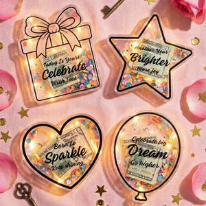 K&ouml;nnte beinhalten: Vier dekorative, transparente Acrylformen mit LED-Lichtern und Konfetti: eine Geschenkbox, ein Stern, ein Herz und ein Ballon. Jede enth&auml;lt einen US-Dollar-Schein und inspirierenden Text. Goldene Schl&uuml;ssel und Sterne sowie rosa Rosenbl&auml;tter umgeben die Formen.