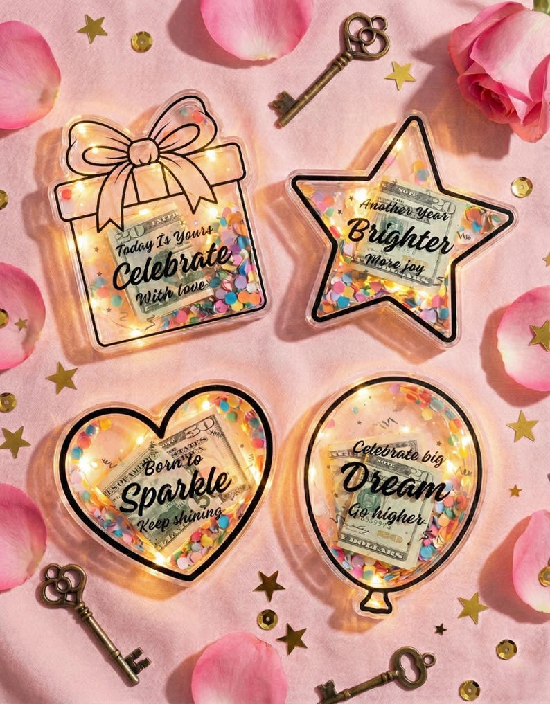 K&ouml;nnte beinhalten: Vier dekorative, transparente Acrylformen mit LED-Lichtern und Konfetti: eine Geschenkbox, ein Stern, ein Herz und ein Ballon. Jede enth&auml;lt einen US-Dollar-Schein und inspirierenden Text. Goldene Schl&uuml;ssel und Sterne sowie rosa Rosenbl&auml;tter umgeben die Formen.