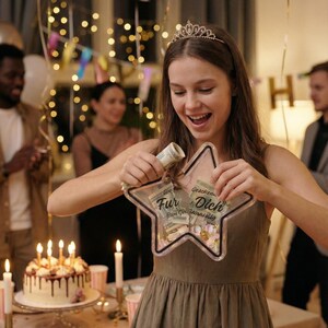 Puede incluir: Una mujer sostiene una caja de regalo en forma de estrella llena de dinero y caramelos. La caja tiene las palabras "Fur Dich". La escena es una fiesta de cumplea&ntilde;os con tarta, globos y velas.