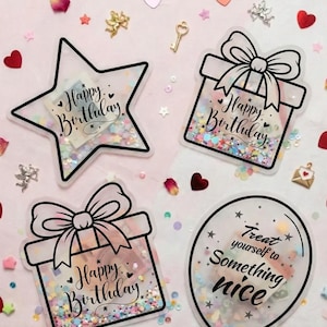Op de afbeelding: Vier transparante acryl verjaardagsdecoraties. Eén is stervormig met de tekst "Happy Birthday", twee zijn in de vorm van een geschenkdoos met strikken en de tekst "Happy Birthday", en één is in de vorm van een ballon met de tekst "Treat yourself to Something nice".