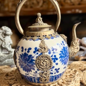 May include: A decorative teapot with a white and blue floral design. The teapot features ornate metal accents, including a handle, spout, and decorative embellishments. The handle is arched, and the spout is curved. The teapot has a lid with a small figure on top.