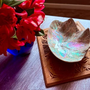 May include: An iridescent, shell-like bowl with a pearlescent finish rests on a square, light brown wooden trivet featuring intricate geometric carvings. The bowl displays a shimmering surface with hints of blue, green, and pink tones.