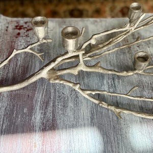 May include: Silver-coloured metal candelabra in a branch design. The candelabra has five candle holders. The decorative piece is made of metal and has a rustic, natural aesthetic. The candelabra is approximately 46 cm long.