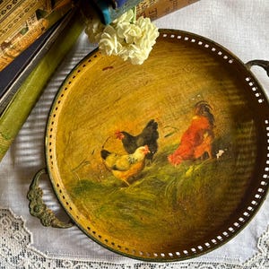 May include: A round metal serving tray with a distressed yellow finish and a painted scene of three chickens in a field. The tray features a perforated rim and two ornate, dark metal handles on opposite sides, showcasing a weathered appearance.