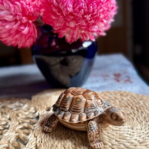 May include: A detailed ceramic turtle figurine with a patterned shell, placed on a woven mat. The turtle is brown and tan. Behind it, a dark blue vase displays bright pink flowers, creating a contrasting backdrop.