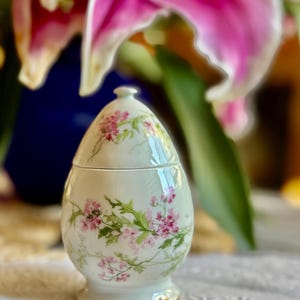 May include: An egg-shaped porcelain trinket box with a lid, decorated with pink floral patterns and green leaves on a cream background. The base has a gold trim. The background features pink lilies and a blue vase.